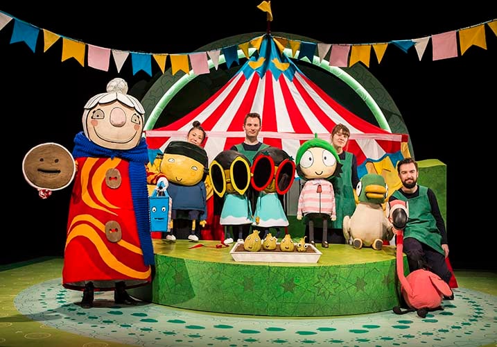 A group of people with puppets and costumes stand in front of a colorful circus tent set, surrounded by playful props and decorations, celebrating Sarah & Ducks Triumphant Theatre Return.